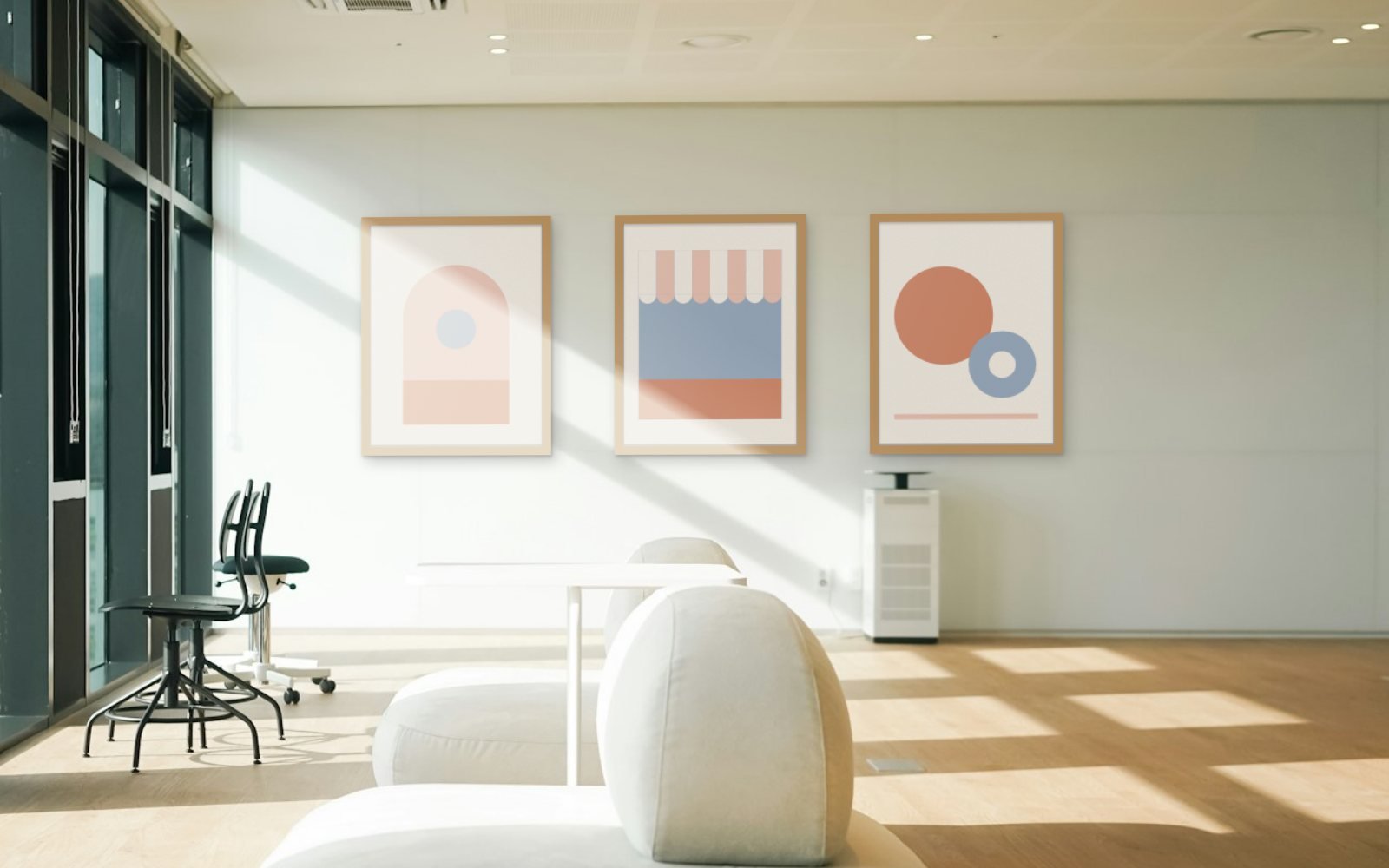 Three Sunroom prints in slim oak frames on a white wall beside floor-to-ceiling windows, a diagonal patch of direct sun falling across the first two prints, with two black office chairs and a white lounge seat below.